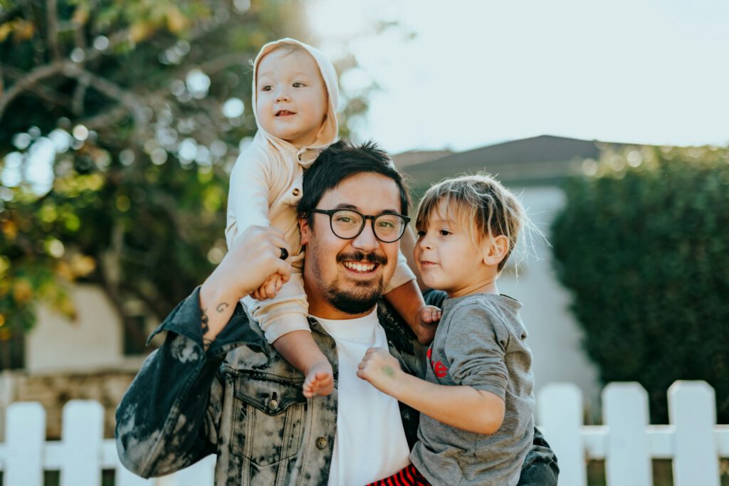 Father smiling outdoors with two young children, one sitting on his shoulders.