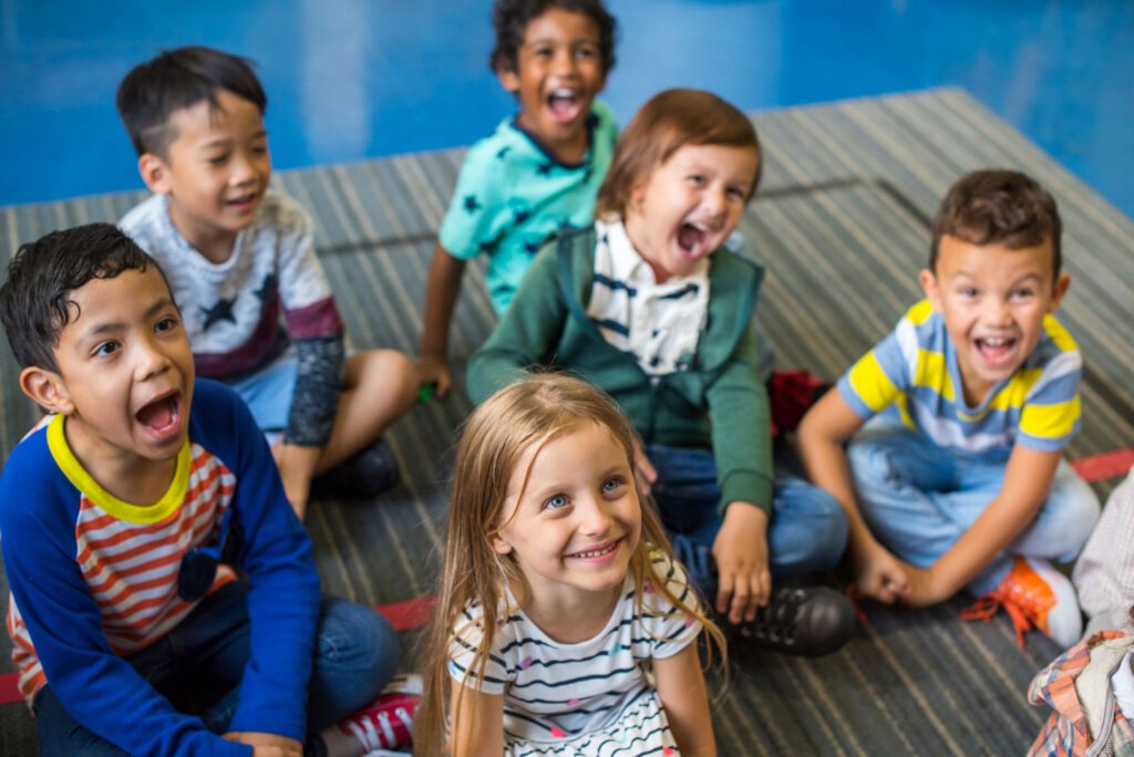 Group of young children sitting on the floor indoors, laughing and smiling together.