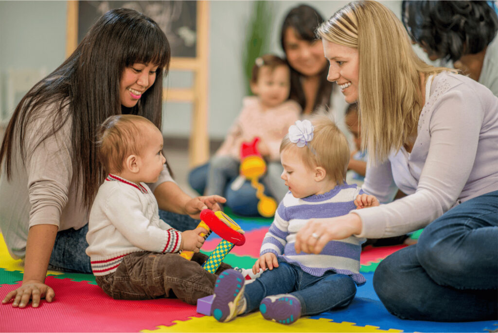 Two women playing with babies on the floor in a group setting.