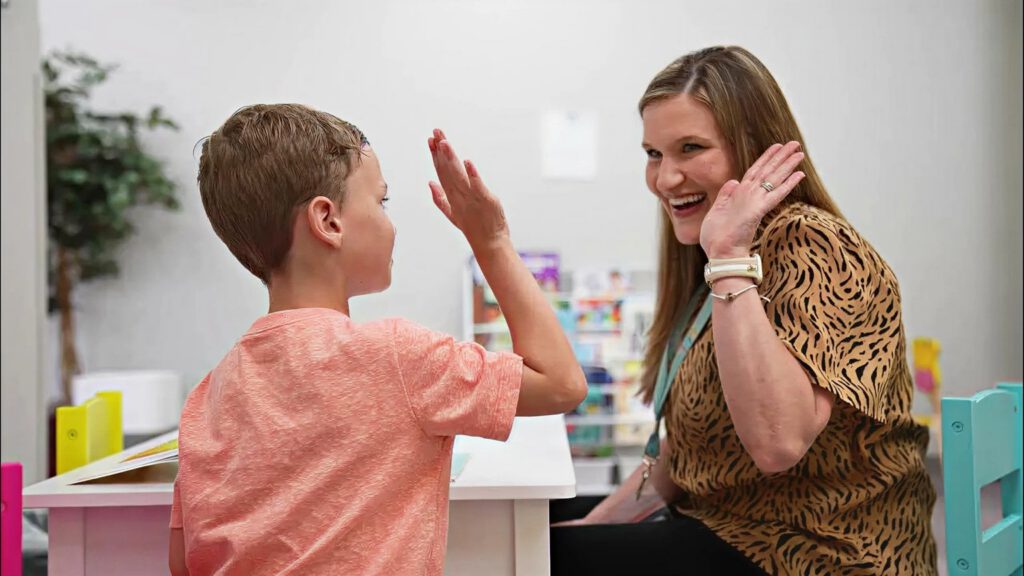 Staff member and child smiling and high-fiving across a table in a classroom or therapy room.