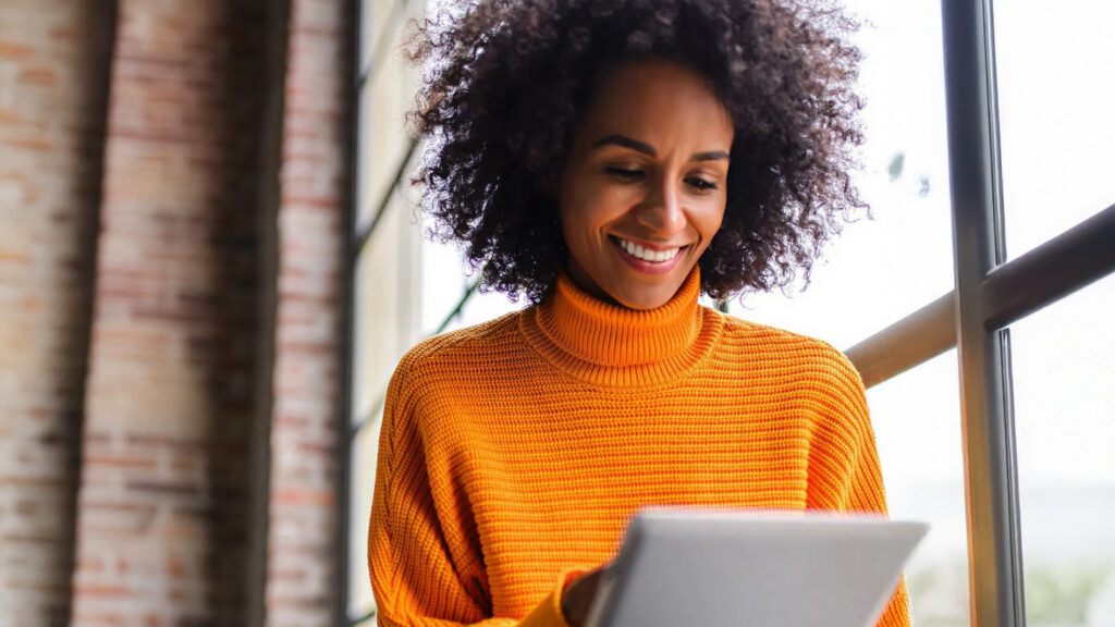 Smiling woman in an orange sweater using a tablet near a window indoors.