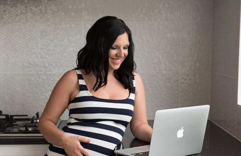 Pregnant woman smiling while using a laptop in a kitchen.