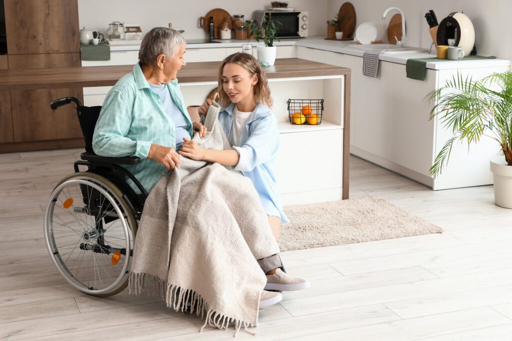 Caregiver comforting an older adult in a wheelchair with a blanket at home.