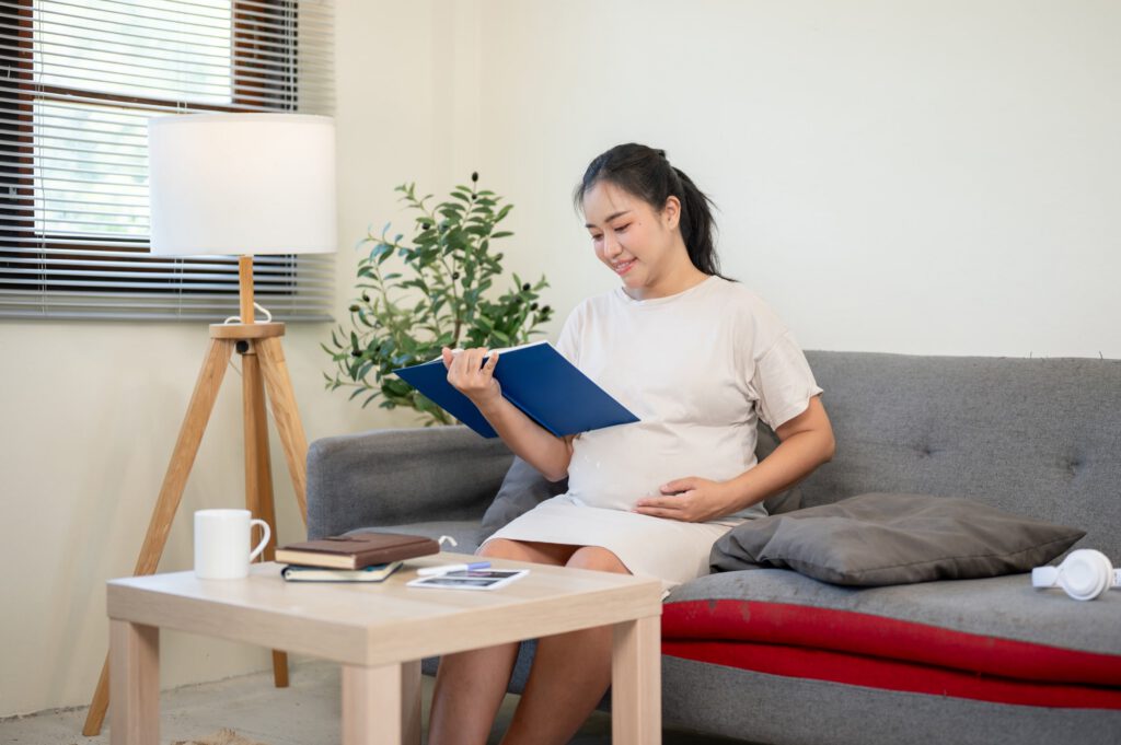 Pregnant woman reads a book while resting on a sofa with one hand on her belly